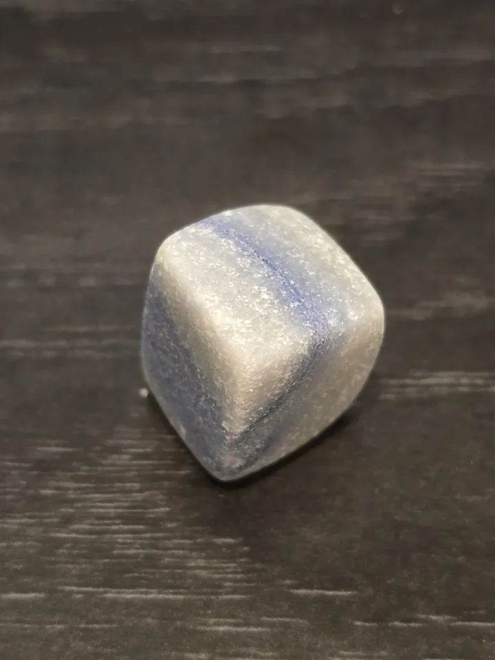 A small, tumbled cubic stone with rounded corners, showing bands of light blue and white with a slightly sugary, crystalline texture, sitting on a dark wooden surface.