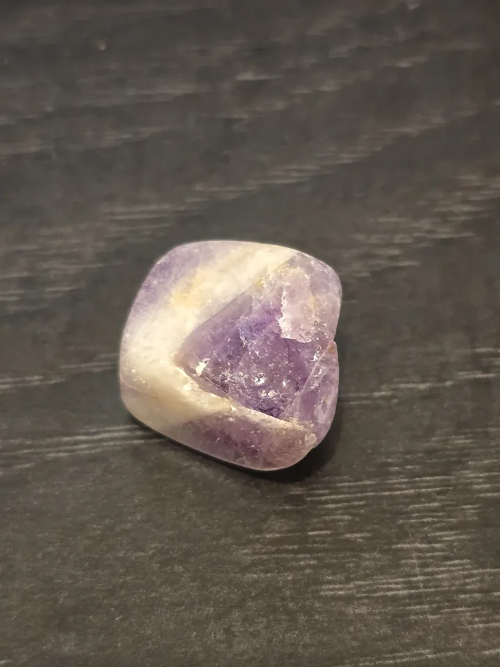 A polished, rounded purple and white stone with V-shaped chevron patterns resting on a dark, wood-textured surface.