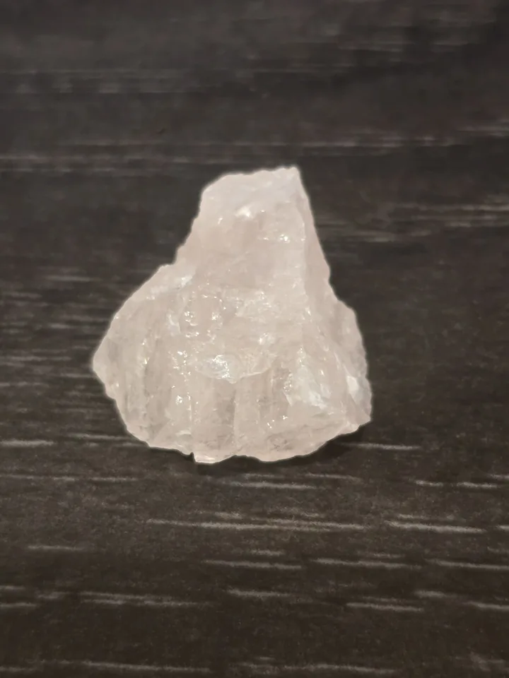 A rough, pale pink piece of rose quartz with a bumpy, crystalline texture, resting on a dark wood grain background.