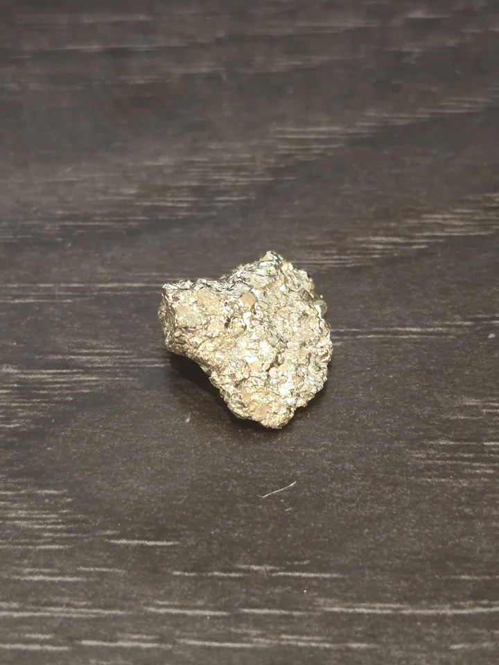A small, rough chunk of metallic, pale brass-yellow mineral with many tiny glittering, blocky crystal faces, resting on a dark wood grain background.