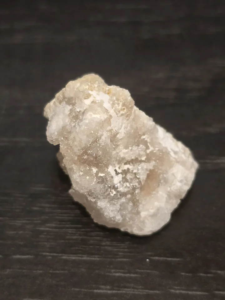A rough, translucent white and light gray mineral specimen with sparkly, crystalline textures, sitting on a dark wooden surface.
