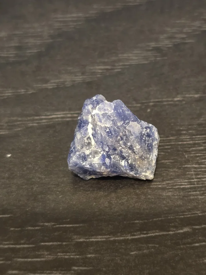 A small, rough, triangular piece of blue mineral with white streaks and a crystalline texture, resting on a dark wooden surface.