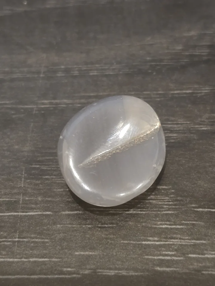 A polished, rounded, translucent light-gray and white stone with a single prominent textured, sandy-looking crack cutting across its center, resting on a dark gray wood-grain surface.