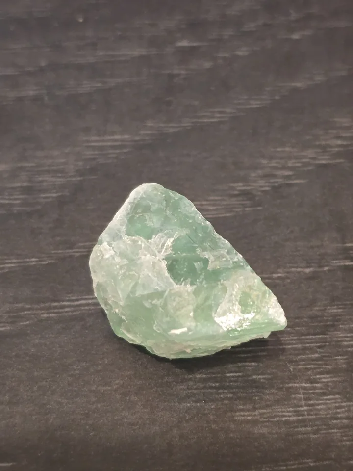 A small, rough chunk of translucent, light green mineral with fractured, glassy surfaces, resting on a dark, textured wood-like background.